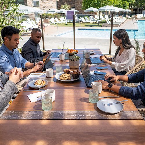 Five people sitting around a table outdoors, working on laptops, with food and drinks on the table, near a pool.