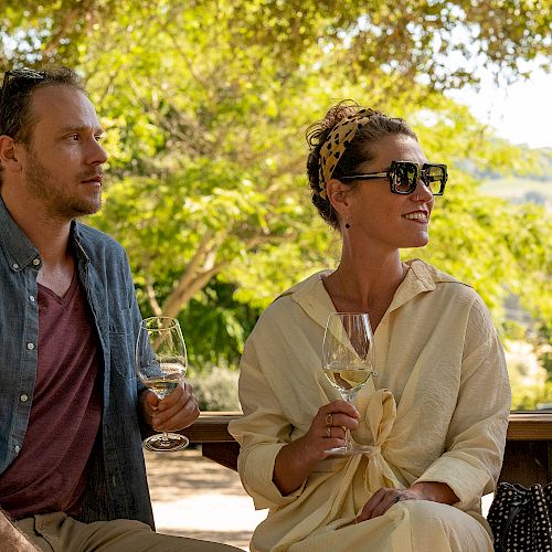 A man and woman are sitting outdoors, enjoying wine. They appear relaxed, dressed casually, and there's a scenic, sunny view in the background.