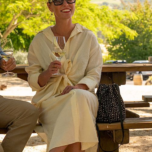 A woman in a light dress sits on a picnic bench, holding a glass of wine and smiling, with trees and hills in the background.