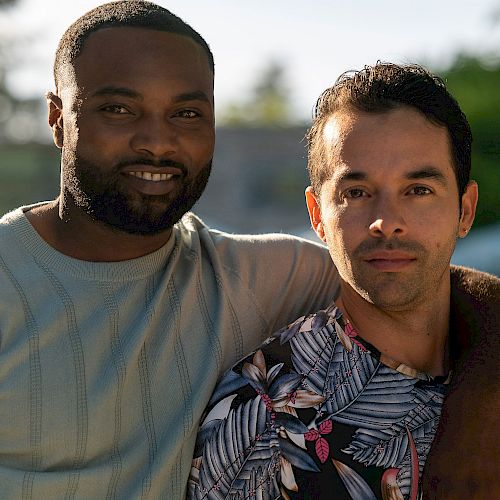 Two people are posing together, one with his arm around the other, smiling outside near greenery and a pool or body of water.