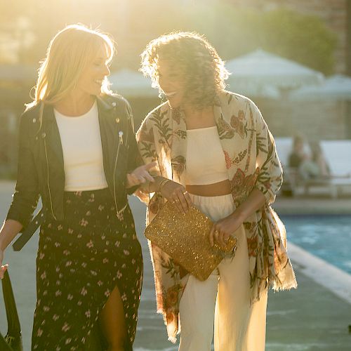 Two women walking beside a pool during sunset, carrying bags and smiling at each other. Sunlight creates a warm backdrop.