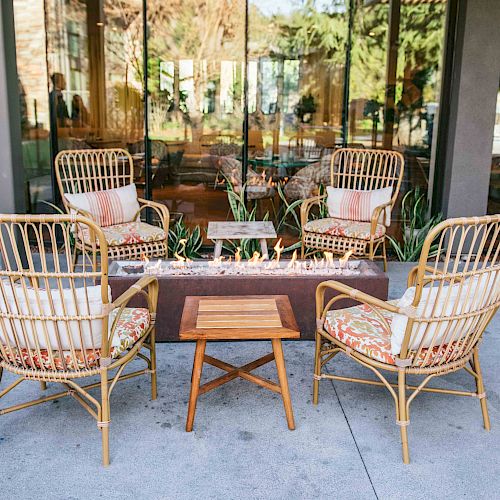 Four wicker chairs with cushions surround a modern fire pit and a small wooden table on a patio, with glass windows and greenery in the background.