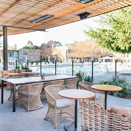 An outdoor seating area with wicker furniture is set next to a swimming pool, surrounded by trees and buildings, under a wooden pergola.