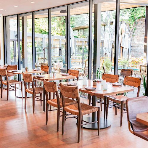 A bright and modern indoor dining area with wooden tables and chairs, large windows, and plants, creating a welcoming atmosphere.