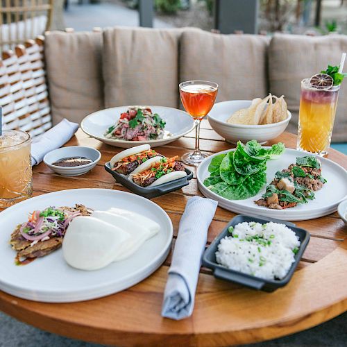 A table with various dishes including tacos, rice, lettuce wraps, steamed buns, and assorted drinks. The setting appears to be outdoors.