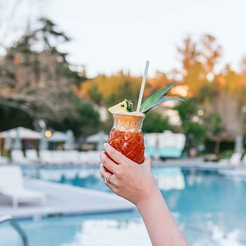 A hand holding a tropical drink with a straw and pineapple slice by a pool, with lounge chairs and greenery in the background.