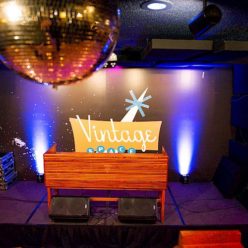 A stage setup with a wooden DJ booth, speakers, and a "Vintage Space" sign in the background. A disco ball hangs from the ceiling.