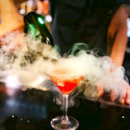 A person is pouring a drink into a red cocktail glass on a bar countertop, with a cloud of smoke or dry ice effect around the glass.