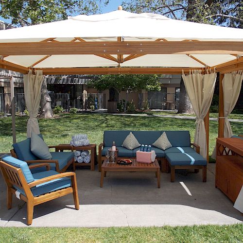 An outdoor cabana with a wooden frame, white canopy, and curtains, furnished with blue cushioned chairs, a loveseat, a coffee table, and a minibar.