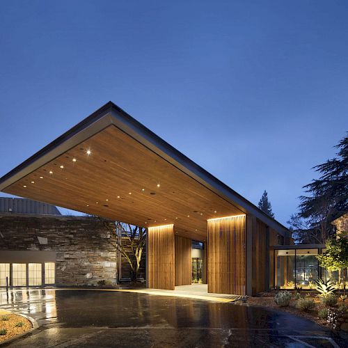 A modern building entrance featuring a large wood-paneled canopy with lights, surrounded by glass windows and a landscaped area, at dusk.