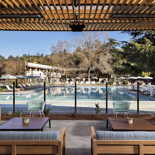 The image shows a luxurious outdoor pool area with lounge seating, tables, and umbrellas, surrounded by trees and modern buildings.