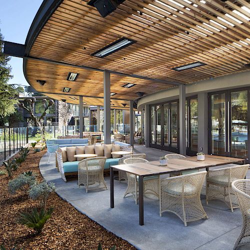 An outdoor dining area with wicker chairs, tables, and cushioned seating under a curved wooden roof looking out towards a landscaped garden and trees.