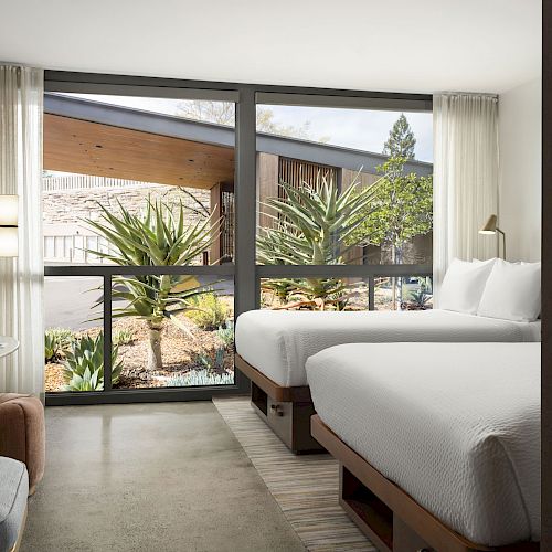 A modern hotel room with two beds, a large window overlooking greenery, a TV, and a seating area with a table and chair, creating a cozy ambiance.