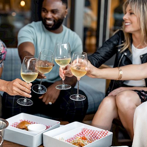 Four people enjoying a meal and drinks, toasting with wine glasses, and smiling at an outdoor setting.