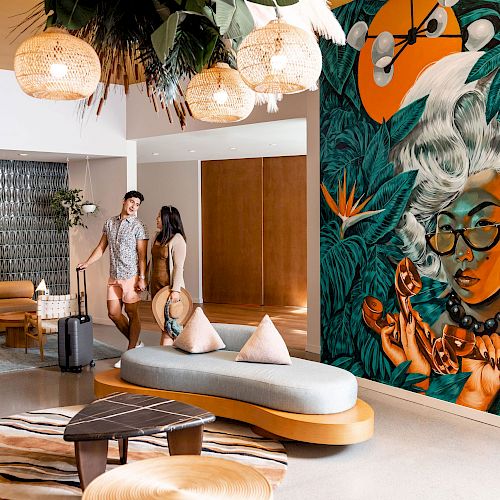 A modern lobby with vibrant wall art, colorful furniture, two people, hanging light fixtures, and tropical decor create a welcoming atmosphere.