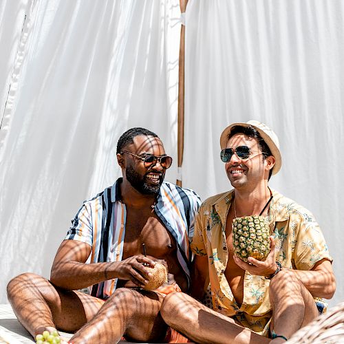 Two men are sitting on a lounge chair, smiling, and enjoying drinks in a sunny, outdoor setting with a white curtain backdrop.