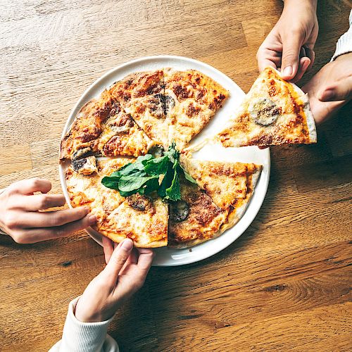 The image shows three hands reaching for slices of pizza topped with greens on a white plate, set on a wooden table.