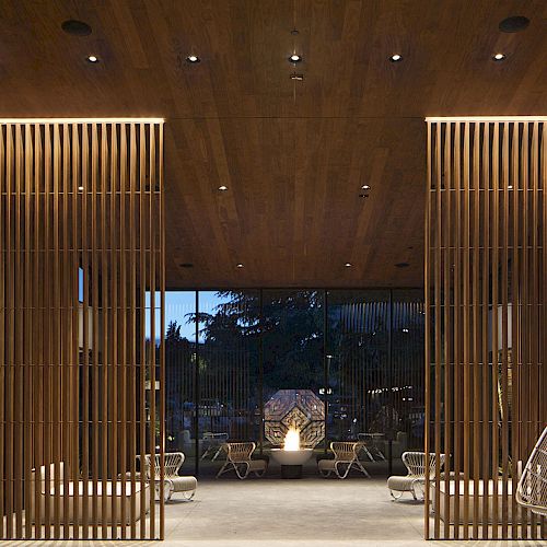 The image shows a modern, open-air lounge area with wooden slat partitions, hanging chairs, and ambient lighting in the evening.