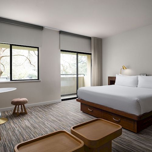 A modern hotel room features a large bed, a wooden table with chairs, a floor lamp, large windows with light curtains, and contemporary decor.