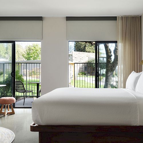 A modern hotel room with a large bed, desk and chair, and sliding doors leading to a balcony with a view of a garden area outside.