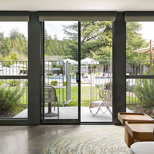 The image shows an interior view of a living room with large sliding glass doors leading to an outdoor pool area with chairs and umbrellas.