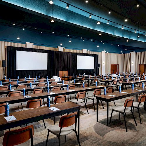 The image shows a conference room setup with rows of tables and chairs facing two large screens and a podium, with water bottles and notepads on the tables.