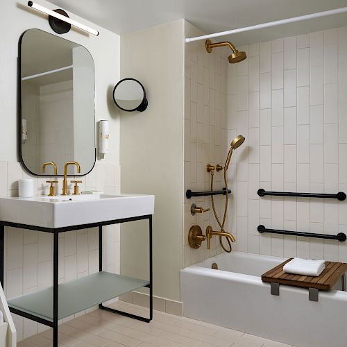 A modern bathroom with a white sink, large mirror, shelves with towels, and a bathtub with gold fixtures and black grab bars.