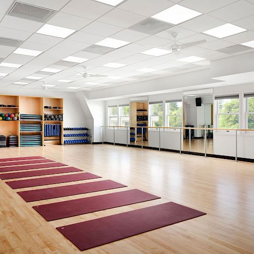 A spacious indoor studio with yoga mats laid out, mirrors on the wall, and shelves holding exercise equipment, featuring natural light from windows.