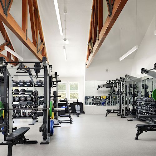 This image shows a brightly lit gym with various weightlifting equipment, benches, and racks, featuring wooden beams on the ceiling for support.