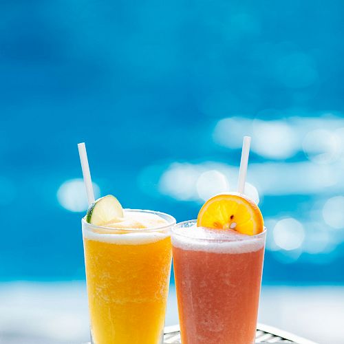 Two refreshing drinks with straws, one orange with a cucumber slice and the other pink with an orange slice, sit on a tray by the pool.