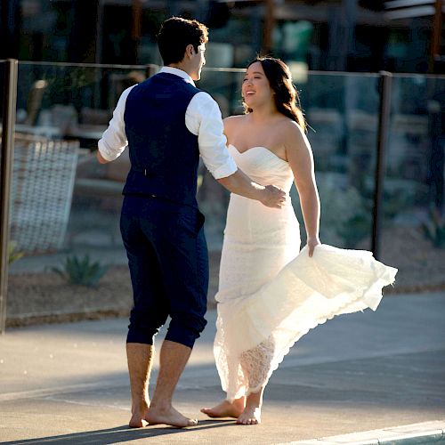 A couple is dancing by a pool, with the woman in a white dress and the man in a vest and rolled-up trousers. They look joyful.