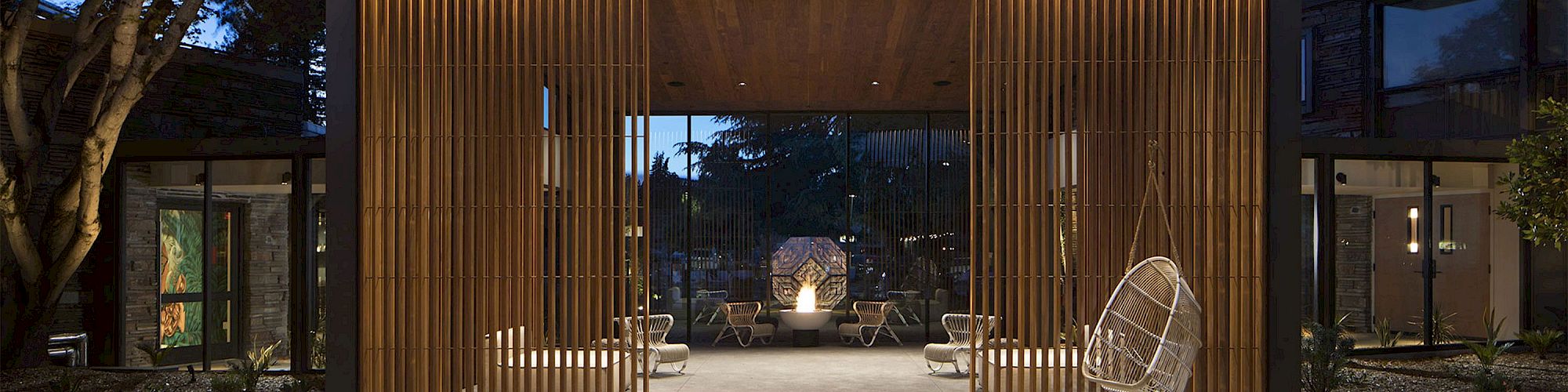 The image shows a modern patio area with wooden vertical slats, hanging chairs, lounge seating, and a central fireplace, all under ambient evening lighting.