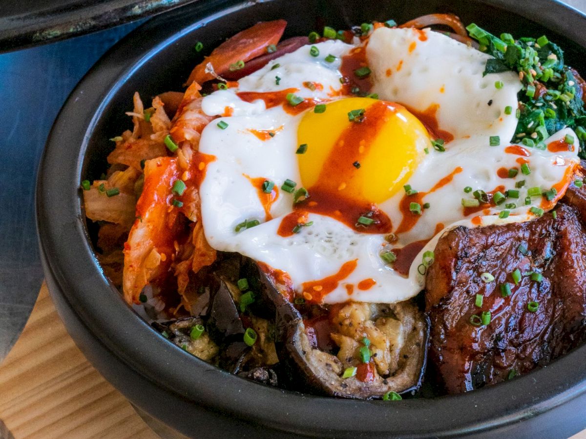 A delicious bowl of bibimbap topped with a sunny-side-up egg, vegetables, marinated beef, and a drizzle of flavorful sauce, served in a stone pot.