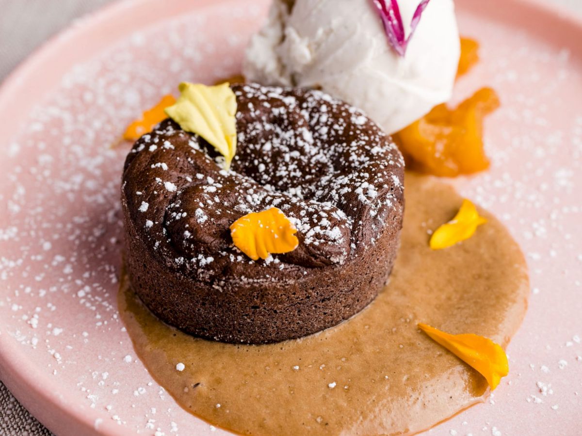 The image shows a chocolate lava cake topped with powdered sugar, surrounded by caramel sauce, with a scoop of vanilla ice cream and flower petals.