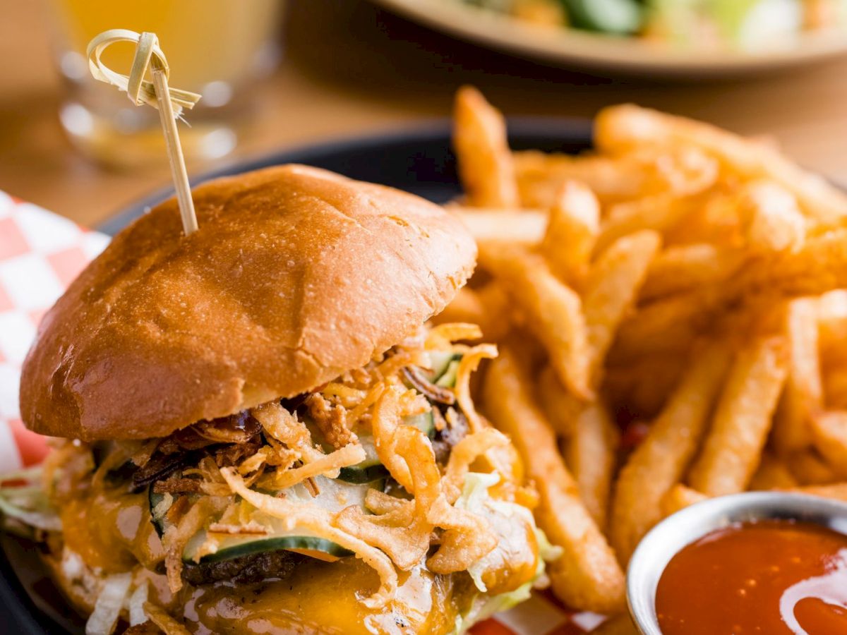 The image depicts a burger with toppings, a side of fries, and a small container of dipping sauce on a checkered paper. A drink and salad are also visible.