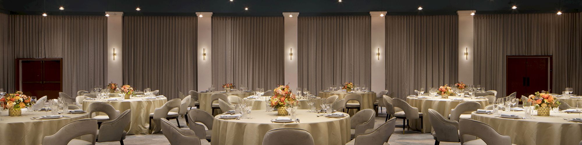 The image shows a banquet hall with round tables set for an event. The room has elegant lighting and floral centerpieces.