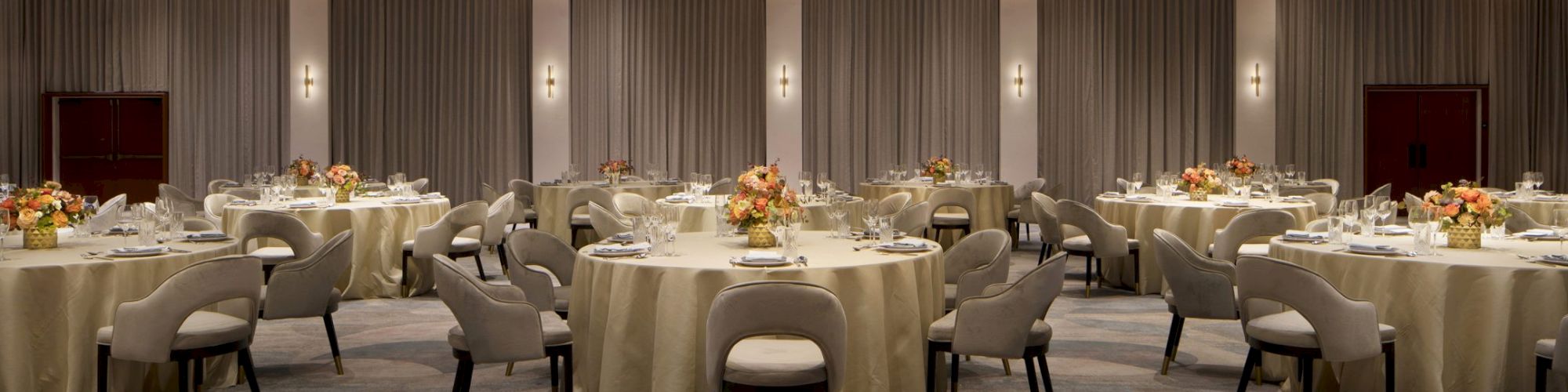 A banquet hall set for an event, featuring round tables with beige tablecloths, elegant chairs, and floral centerpieces.