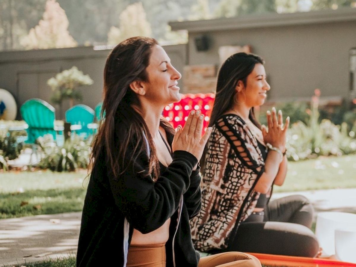 Two individuals are meditating on yoga mats in an outdoor setting with greenery and buildings in the background.