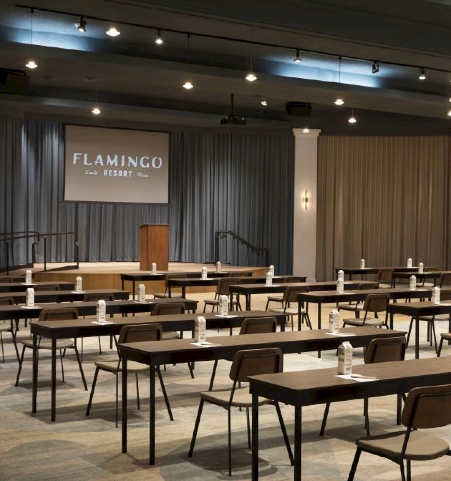 The image shows a conference room at Flamingo Resort with rows of tables and chairs, a stage, a podium, and a projection screen.