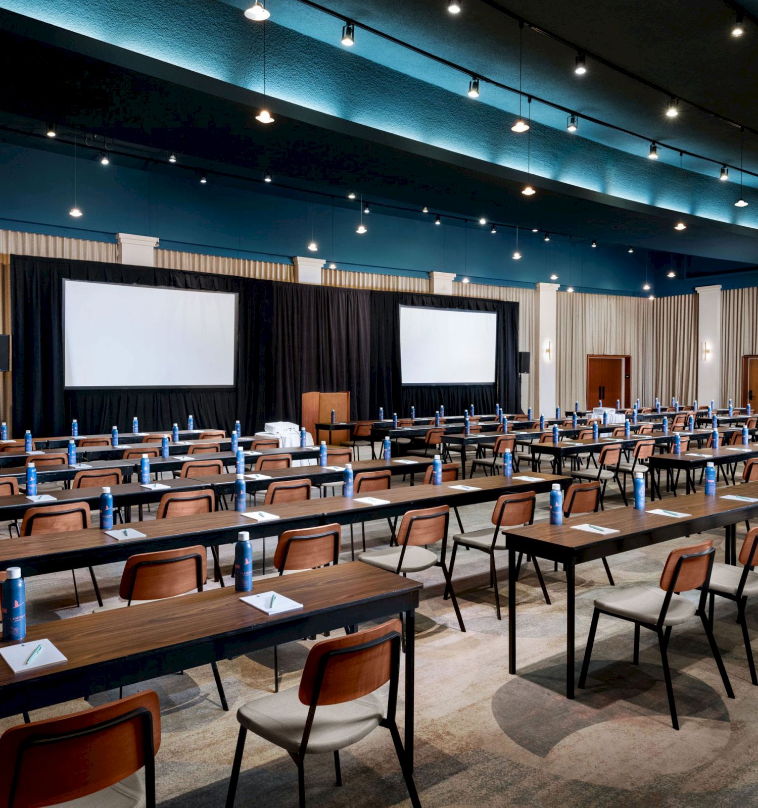 A conference room with rows of tables and chairs facing two large screens, each setup with water bottles and notepads on the tables.