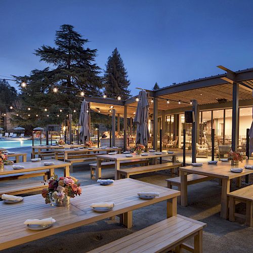 An outdoor dining area with wooden tables and benches, string lights, and a serene evening setting with trees and a pool in the background.