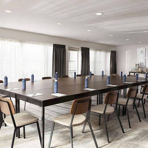 A conference room with a large table set for a meeting, having water bottles, notepads, and chairs arranged around. Refreshment area in the background.