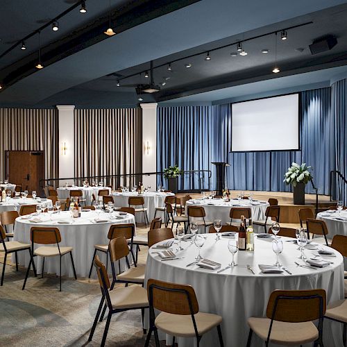 The image shows a banquet hall with round tables set for an event, featuring a stage, blue curtains, and a projector screen, ending the sentence.