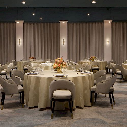 The image shows a banquet hall with round tables set for an event. The tables are decorated with flowers and chairs are arranged around them.