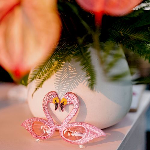 A pair of pink flamingo-themed glasses is placed in front of a white vase with green fern leaves and pink flowers.