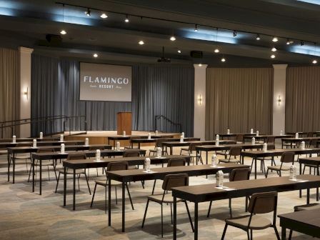 An empty conference room with rows of tables and chairs facing a stage with a podium and a screen displaying 