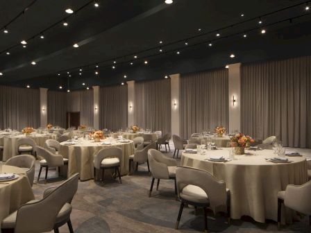 The image shows a banquet hall with round tables covered in beige tablecloths, floral centerpieces, and elegant chairs arranged neatly.