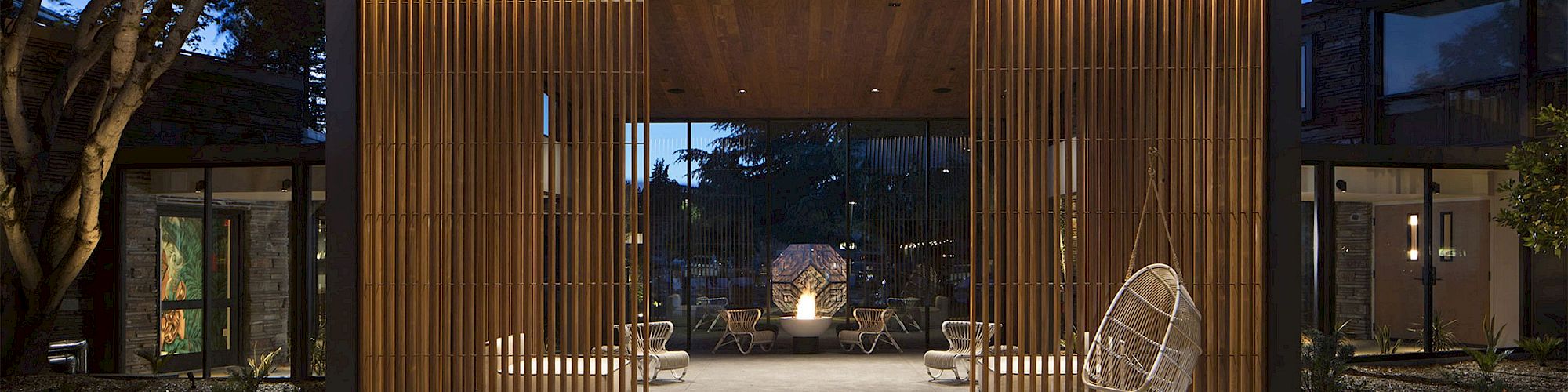 A modern, open-space lobby with wooden slats, hanging chairs, and outdoor seating areas arranged around a central fireplace, seen from outside.