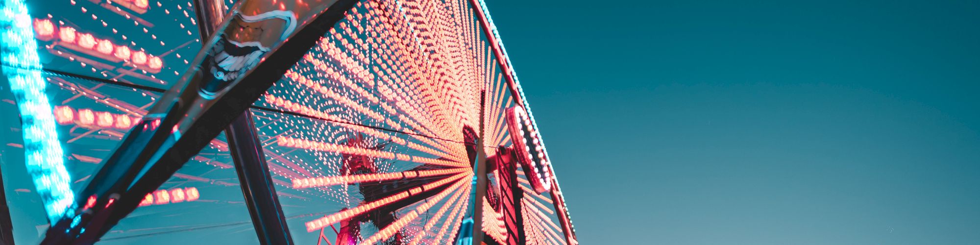 The image shows a close-up of a brightly lit Ferris wheel against a clear sky, with colorful lights creating a vibrant and dynamic pattern.