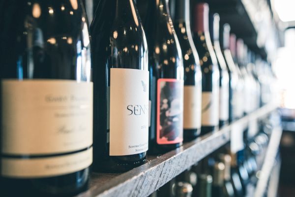 This image shows a row of wine bottles lined up on a shelf, each with distinct labels, in what appears to be a wine shop or bar setting.